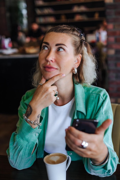 Beautiful stylish woman in green shirt sitting in loft cafe drinking cappuccino and checking her social media at phone