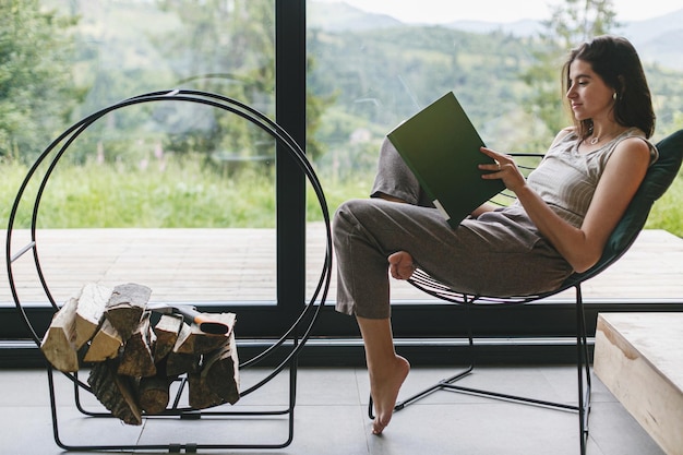 Photo beautiful stylish woman reading book sitting on chair at fireplace on background of mountain hills