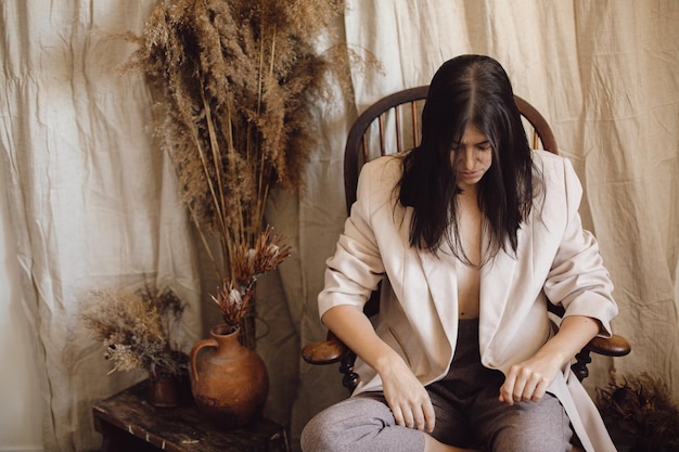 Photo beautiful stylish woman in suit sitting on retro wooden chair in bohemian style room with dry grass