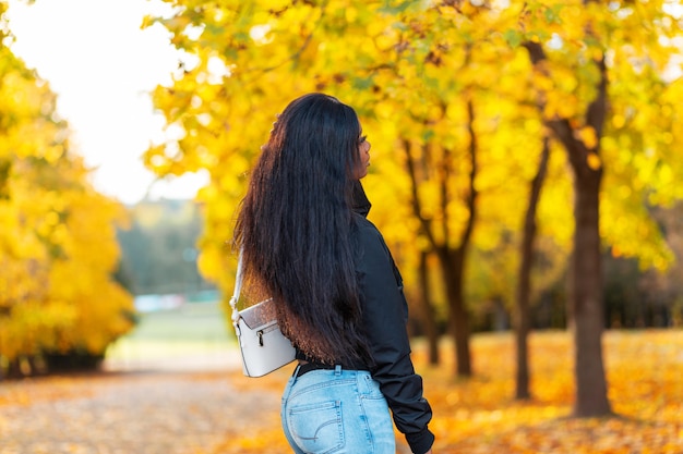 Красивая стильная молодая африканская модель женщины с длинными волосами в модной черной куртке с джинсами и сумкой гуляет в осеннем парке с ярко-желтой листвой падения. Женский повседневный стиль и красота