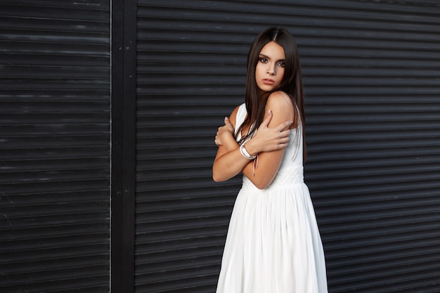 Bella ragazza alla moda con un viso carino in un abito bianco vicino a un muro di metallo scuro