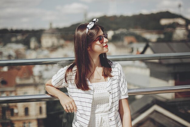 Beautiful stylish young woman in sunglasses retro dress and headband relaxing on roof in european city street Happy hipster girl smiling and enjoying summer time in Lviv Copy space