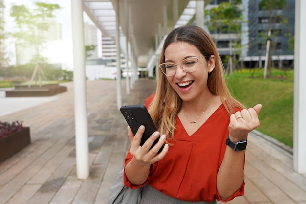 Beautiful successful Brazilian female excited raised hand rejoicing with smartphone outside Young woman watching at mobile phone celebrating good news from work in the morning