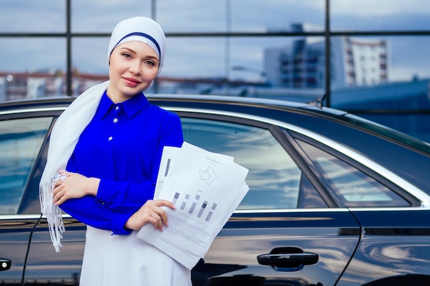 Beautiful and successful european muslim business woman entrepreneur in stylish hijab and turban holding paper skyscraper office windows background on street standing near her black car