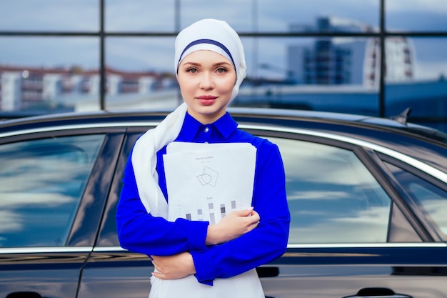 Beautiful and successful european muslim business woman entrepreneur in stylish hijab and turban holding paper skyscraper office windows background on street standing near her black car