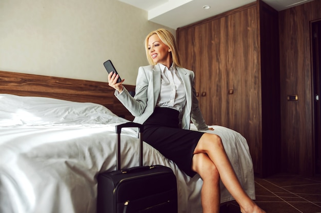 Photo beautiful and successful woman in formal clothes is sitting on the bed in a hotel room and using a phone