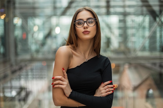 Beautiful successful young business lady with fashion glasses in a stylish black top with one shoulder in a glass office building