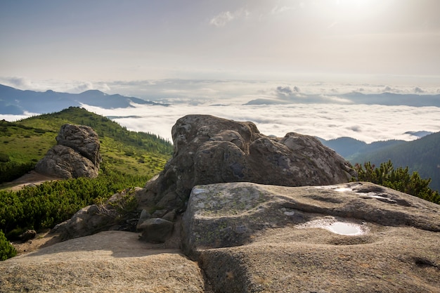 Beautiful summer mountain panorama at dawn. Huge boulder with rain water puddle on green rocky mountain top on  of foggy valley filled with white puffy clouds and clear blue copy space sky.