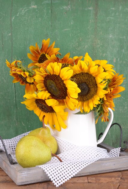 Beautiful sunflowers in pitcher with pears on table on wooden background