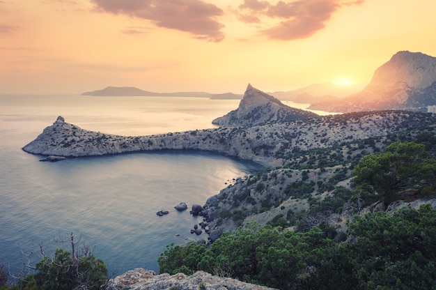 Beautiful sunrise in mountains at the sea. Amazing summer landscape with rocks, mountain forest, colorful bright orange sky with sun, blue water in the morning. Seascape at dawn. Nature background