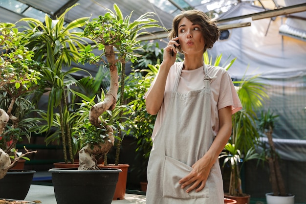 beautiful surprised woman gardener standing over plants in greenhouse talking by mobile phone.