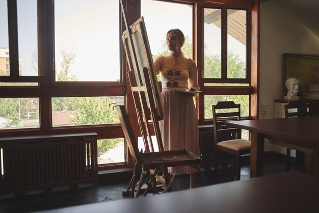 Bella giovane artista di talento dipinge un'immagine con pennello e olio su un cavalletto con tavolozza in mano in un elegante studio d'arte creativa