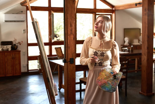 美しい才能のある若い女性アーティストは、スタイリッシュなクリエイティブアートスタジオでパレットを手にイーゼルにブラシと油で絵を描く