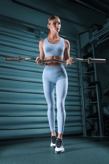 Beautiful tall blonde posing in the gym with a barbell in her hands against the background of the wall bar. The concept of sports, fitness, aerobics, bodybuilding, stretching. Front view.