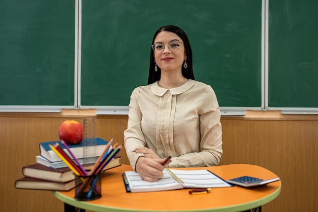 Beautiful teacher with glasses sits at a desk with book, pencil notepad apple against blackboard, classroom