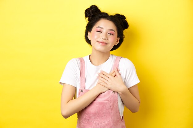 Beautiful teen asian girl in love, holding hands on heart and smiling touched, looking with affection, standing on yellow.