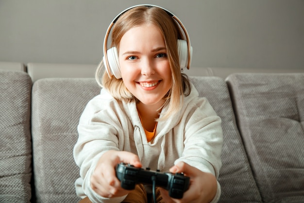 Beautiful teen girl blonde playing video console game using game controller joystick gamepad sitting on couch sofa. Happy young woman player wins in game console while playing video game at home party