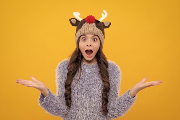 Beautiful teen girl in a winter hat and a warm sweater a child on a yellow isolated background excited face cheerful emotions of teenager girl