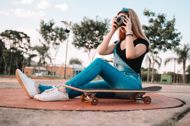 Beautiful teenage girl sitting on a skateboard outdoors taking pictures with a camera.