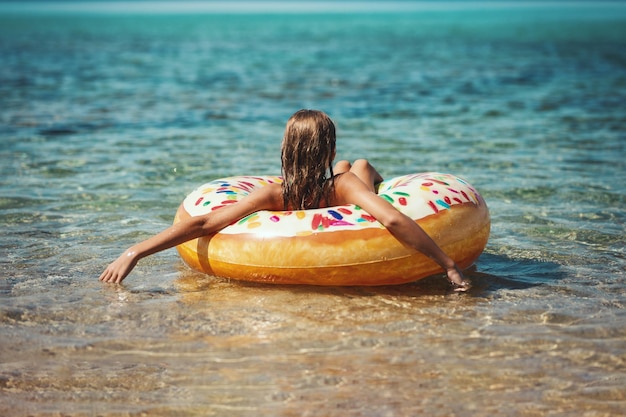 美しい10代の少女は、海の水で膨脹可能なリングドーナツで泳ぎ、休暇で楽しんでいます。
