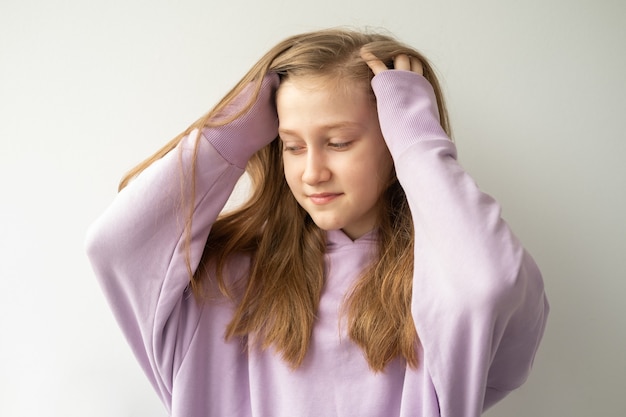 Beautiful teenage girl with long hair in purple huddyhoodie touching her hair standing against white wall