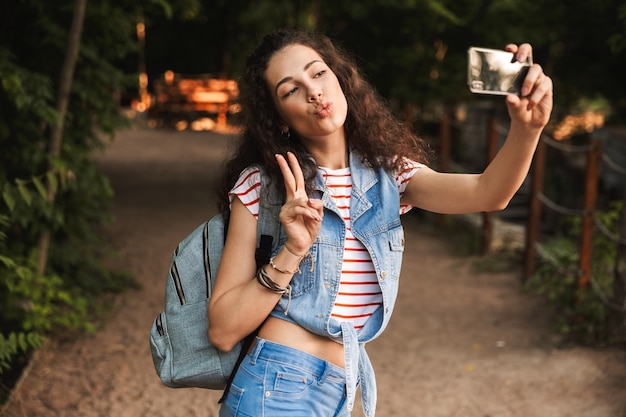 beautiful teenage woman with backpack, showing peace sign and taking selfie on smartphone while walking outdoor