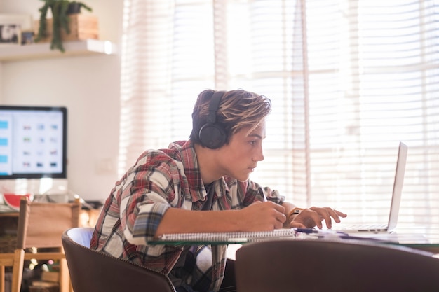 Beautiful teenager focus for his studies doing homework at home on the table with laptop or computer - headphones on the table - indor lifestyle concept - guy writing and reading