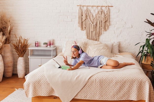 Beautiful teenager girl communicates online by phone while lying on the bed at home. Child in headphones learn online while lying on the bed. Distance learning concept
