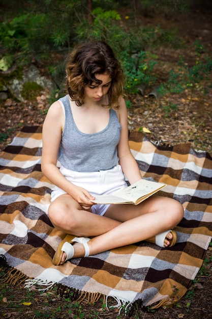 写真 森の緑の草の上に毛布の上に横たわって本を読んでいる美しいティーンエイジャーの女の子