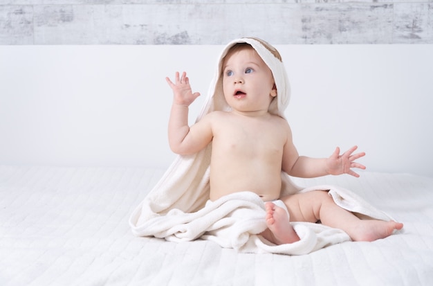 Beautiful ten month baby girl sitting on bed indoors in light modern interior with towel after bath. Baby care concept.