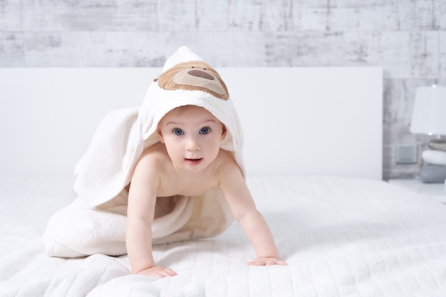 Beautiful ten month baby girl sitting on bed indoors in light modern interior with towel after bath. Baby care concept.