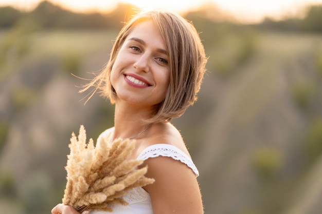 Beautiful tender girl in a white sundress walks at sunset in a field with a spikelet bouquet.