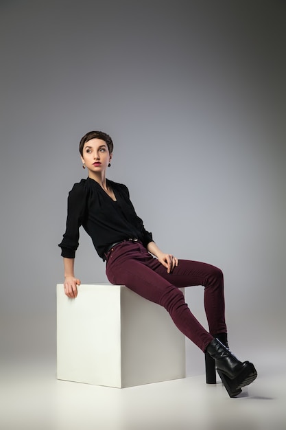 Beautiful thin brunette woman  sitting on cube over gray background