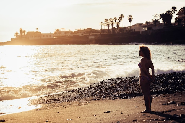 自然と触れ合う休暇と自由なライフスタイルを楽しむために海の波の近くの海岸に立っている美しい薄い女性白人。地平線と日没時間。砂の上の足と夏の幸せ