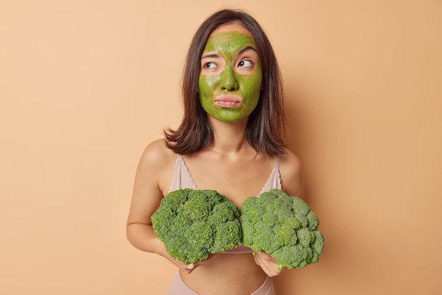 Beautiful thoughtful asian woman with dark hair applies
nourishing natural vegetable mask on face holds broccoli looks away
wears top takes care of skin isolated over beige studio
background