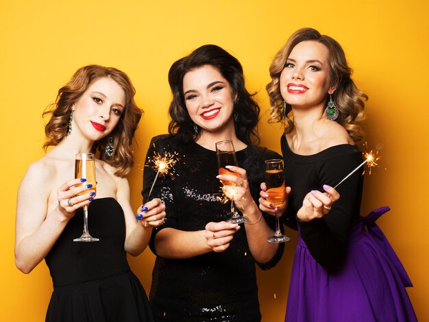 Beautiful three young women with a champagne glasses and sparklers over yellow background