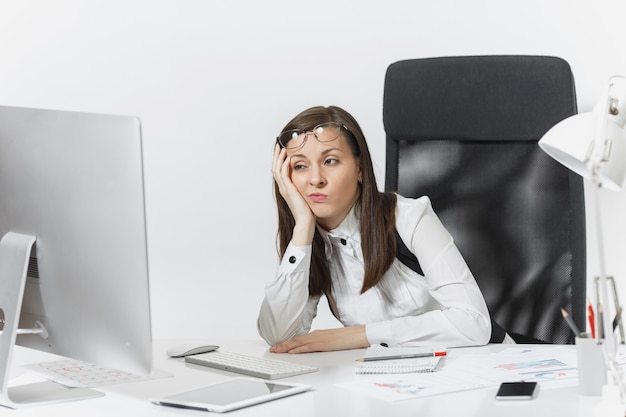 La bella donna d'affari stanca perplessa e stressata dai capelli castani in giacca e occhiali seduta alla scrivania, lavorando al computer contemporaneo con documenti in ufficio leggero