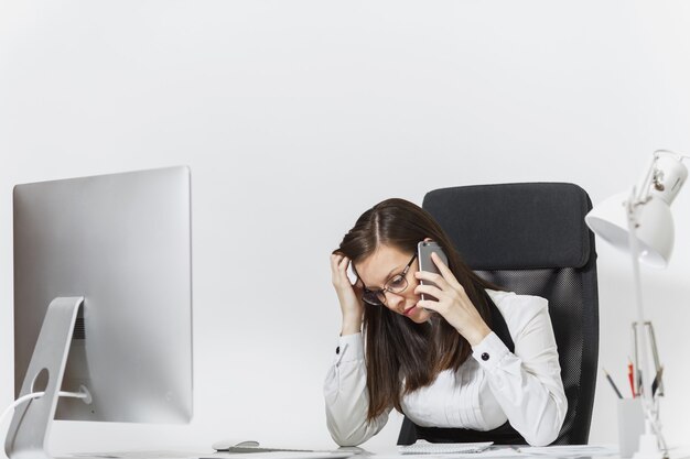 Beautiful tired and stress business woman in suit sitting at the desk, working at modern computer with documents in light office, talking on mobile phone, resolving issues