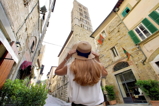 美しい観光客の女の子は、アレッツォ トスカーナ イタリア低角度の中世の町を歩いて帽子を保持します。