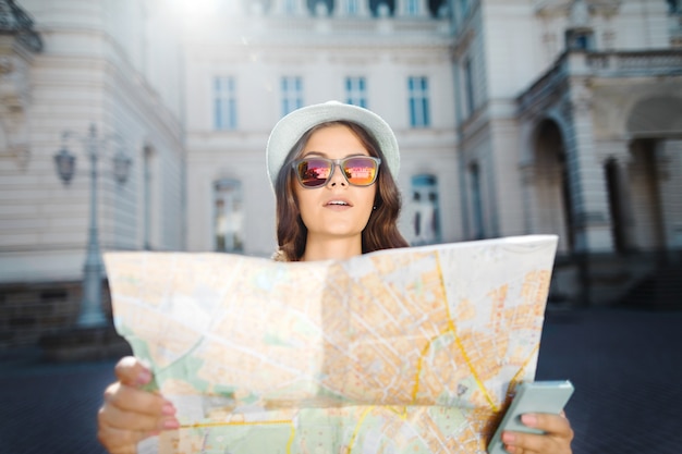 Photo beautiful tourist girl with curly hair wearing hat and sun glasses, holding map and mobile phone at old european city, traveling, europe.