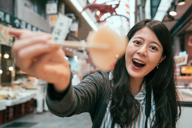 写真 箸でカメラに刺身を見せる美しい観光客。日本食を食べるアジアの女の子の旅行者に焦点を当てた写真。 jpの大阪の通りの新鮮なシーフード。