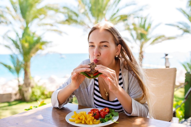 Beautiful Travel Woman with Fastfood Burgers Coffee and Juice Drinks at Outdoor Cafe with Blue Sea and White Sand on Background Smiling Woman with Long Hair at Outside Restaurant in Sunny day