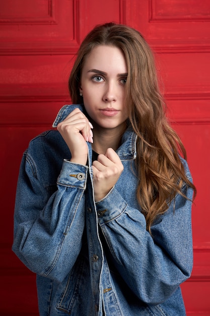 Foto bella ragazza alla moda o modello con i capelli lunghi che indossa in una giacca di jeans in piedi sul muro rosso