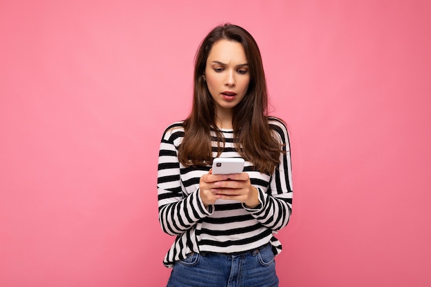 Beautiful upset thoughtful young brunette woman using mobile phone communicating via texting message wearing sweater isolated on wall .