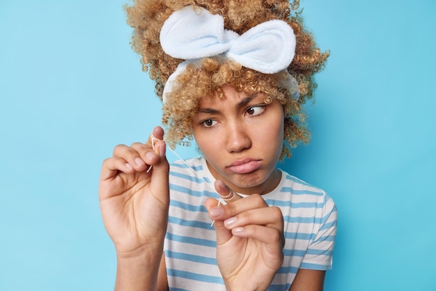 Foto bella donna sconvolta con capelli biondi ricci guarda attentamente il filo interdentale andando a pulire i denti e rimuovere il cibo indossa una maglietta a righe casual con fascia isolata su sfondo blu per studio