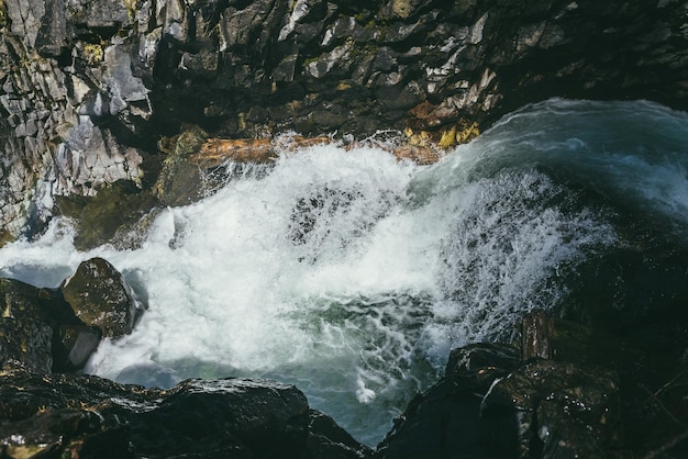 Beautiful view from above to turquoise turbulent mountain river among rocks in narrow gorge in sunshine. Atmospheric mountain landscape with wet mossy stones in powerful mountain river in sunlight.