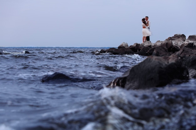 岩の多いビーチで美しい結婚式のカップル。日没で手をつないで、屋外を抱いてスニーカーでスタイリッシュな新婚夫婦。海岸に立っている新郎新婦。自然に恵まれた日。ロマンチックなデート