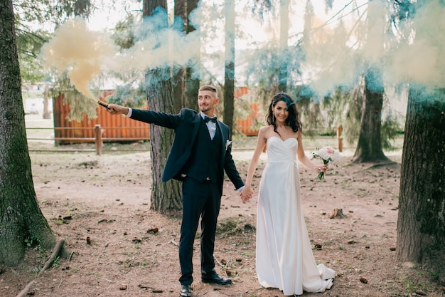 Beautiful wedding couple standing together at nature and holding colorful smoke.