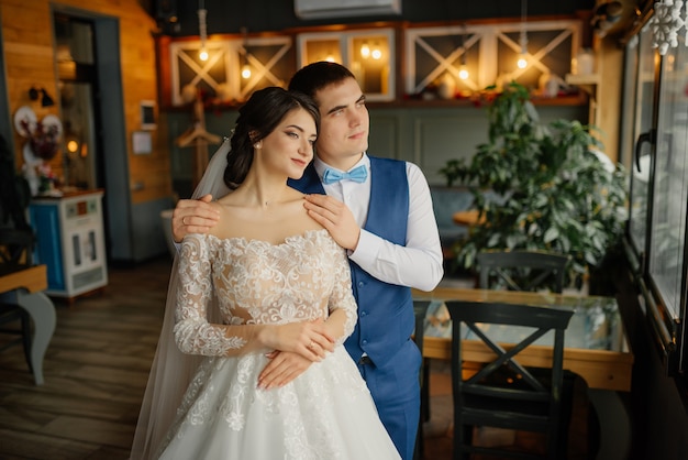 Beautiful wedding, husband and wife, bride and groom standing in loft interior near window. Newlyweds couple in love. Groom hug the bride by the shoulders. Bride hold a wedding bouquet in hands.