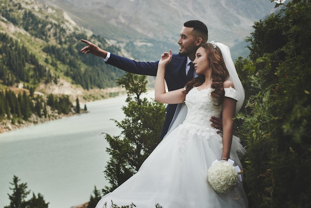 Beautiful wedding photo on mountain lake. Happy Asian couple in love, bride in white dress and groom in suit are photographed against background of the Kazakh landscape